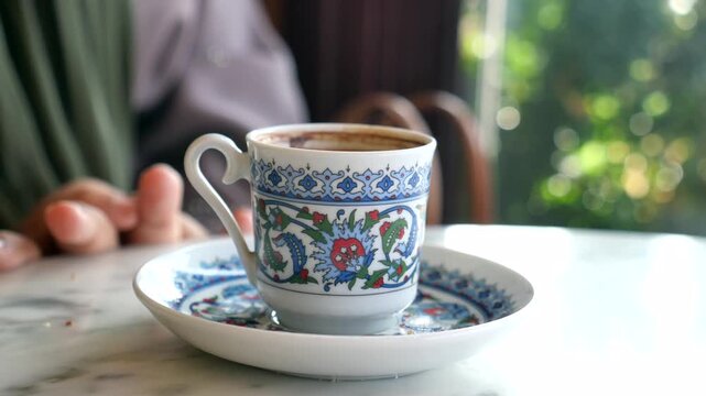 A close up slow motion video of a person picking up a decorative cup of turkish coffee from a saucer on a white marble table