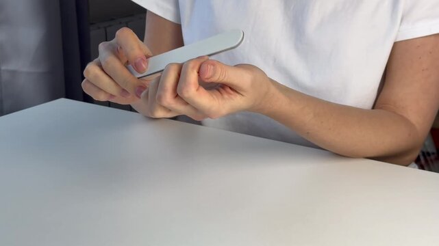 A European woman sits at a table and files her nails. Home beauty treatments. Self-care. Medium shot. Home manicure.