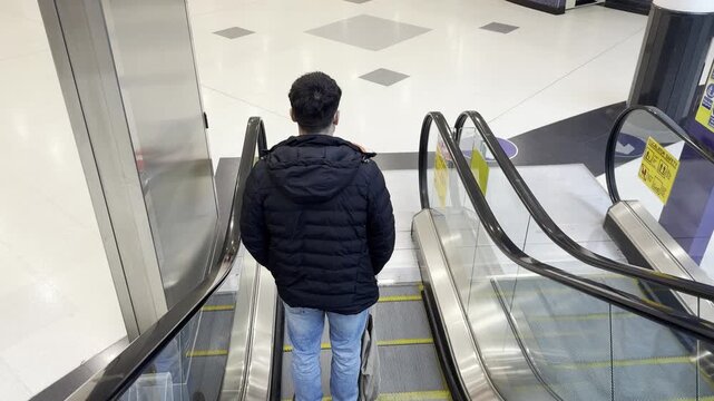 A man descends an escalator in a shopping mall. View from above. The man is photographed from behind.