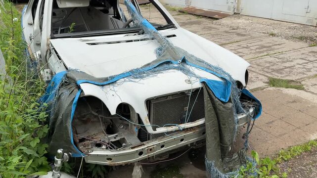 Dismantled wrecked sedan front end exposed, blue tarp draped over mangled grille, missing headlights, twisted bumper and exposed radiator, weeds and grass encroaching on abandoned vehicle.