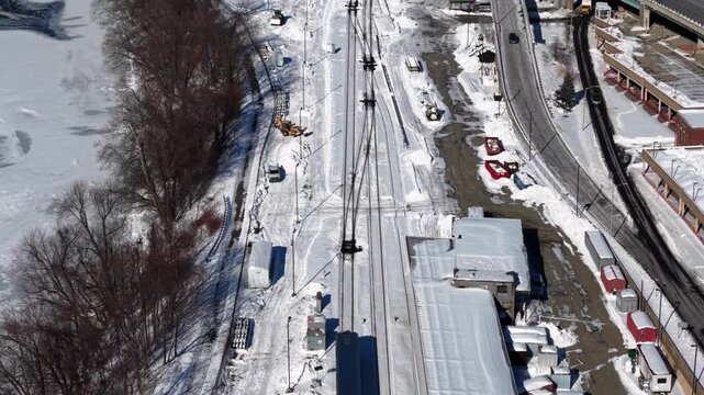 Aerial view of freight train moving through snowy industrial area in Springfield, Massachusetts, with rail tracks, warehouse rooftops and winter landscape under clear daylight. Top down.
