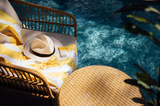 Woven rattan chair with yellow striped towel, sun hat, and sunglasses beside a sparkling blue swimming pool in a tropical outdoor setting