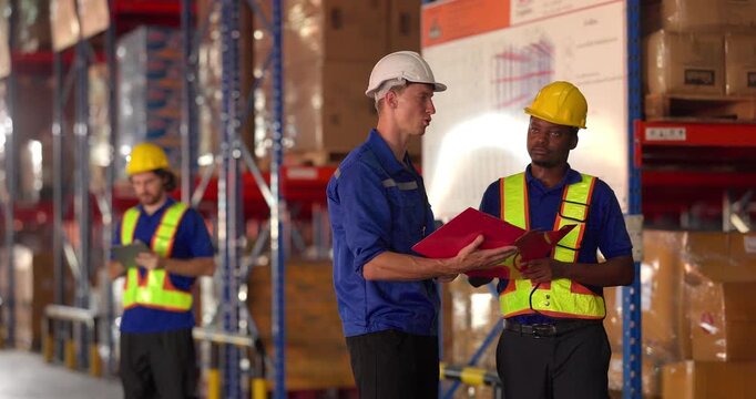 Three warehouse workers in safety vests and helmets coordinating logistics with a tablet and clipboard beside a forklift and pallet of boxes in an industrial storage facility.
