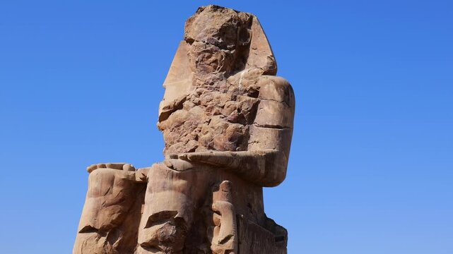 Giant stone colossi at Memnon archaeological site in Luxor valley, ancient Egyptian pharaoh statues with throne carvings against dramatic desert cliffs