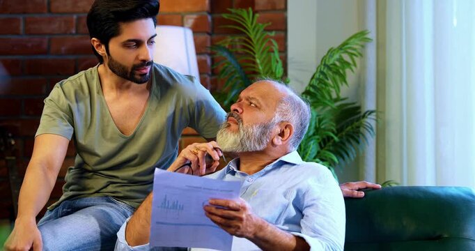 Indian retired father and young son discussing finance while reviewing financial report, profit and loss statement and business growth documents together on sofa in modern home interior