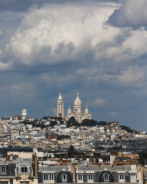 View of the Sacre-Coeur Basilica atop Montmartre hill, a beacon against the dramatic sky, overlooking the Parisian rooftops, Paris, Ile-de-France, France.
