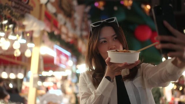 Beautiful young asian woman traveler using smartphone taking selfie with noodle street food at a vibrant night market with local cuisine experience and nightlife atmosphere in travel urban lifestyle.
