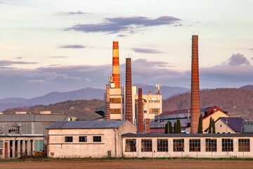 Fototapeta premium Glass factory with industrial buildings and tall chimneys. In the background are blurred mountains and evening sky with clouds.