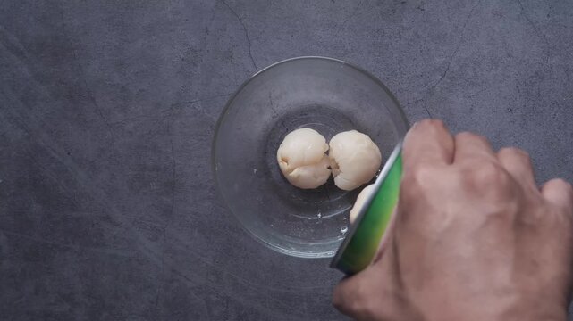Overhead view of fresh lychee fruit being poured from a can into a clear glass bowl, showcasing the juicy texture and natural color of the fruit with a dynamic motion,
