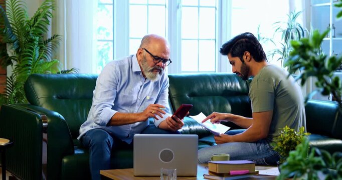 Indian retired father and young son working together on laptop and smartphone at home, reviewing financial report, share trading data and online business documents while discussing profit and loss