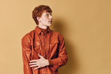 Young man with red hair wearing brown leather jacket and turtleneck looking sideways on beige background. Profile portrait with casual style and fashion expression indoors. © SHOTPRIME STUDIO
