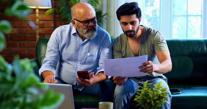 Indian retired father and young son working together on laptop and smartphone at home, reviewing financial report, share trading data and online business documents while discussing profit and loss