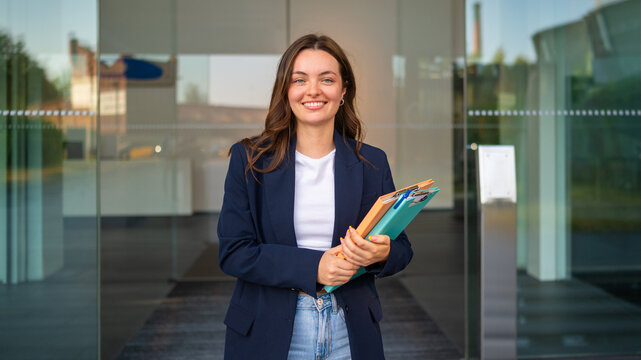 Confident young businesswoman holding folders smiling at office entrance