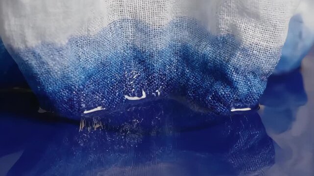 White fabric slowly dipping into blue liquid in slow motion. Macro close-up of absorption and dyeing process. Textile and chemistry concept