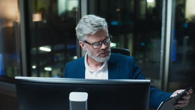 Male professional multitasking at desk during night shift. Caucasian businessman wearing glasses checking notepad while watching at desktop monitor. Analyzing data and comparing information.