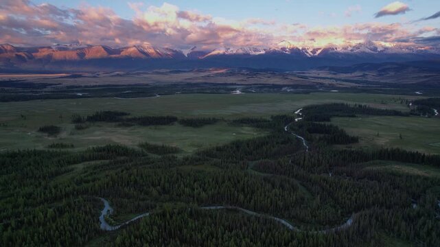 Aerial reveal pulling back and tilting up over winding river and dense forest toward snowcapped Altay mountain range bathed in warm sunset light for nature background.