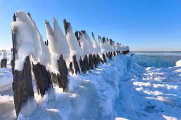 Frozen coastal pier reaching into silence &mdash; ice-covered wooden posts, deep blue sky and long winter shadows creating a serene northern seascape of cold light and quiet strength.