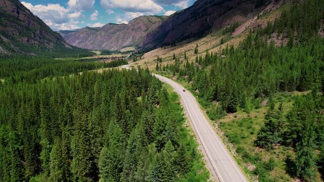 Aerial tracking shot of red car driving along winding Altai mountain road through green forest, trip for travel and adventure.
