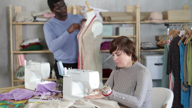 Woman tailor sews clothes in sewing machine. Worker uses sewing machine to create clothes for clients. African male companion work in background with mannequin