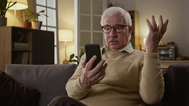 Medium zoom shot of senior Caucasian man with gray hair relaxing at home with smartphone, receiving money withdrawal alerts from scam attack, lifting up glasses and staring at shock and disbelief