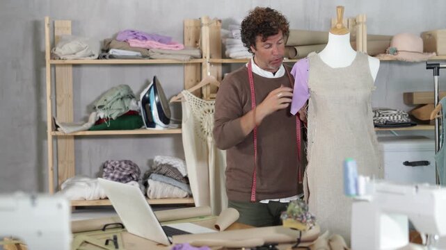 Male fashion designer communicates with a client via video call from a sewing workshop and shows a sample of clothes on a mannequin. Tailor shows the customer the quality of the sewn clothes. High