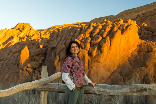 A smiling woman in a patterned shawl stands by a wooden fence with a rock formation behind her called Monkey fingers. Dades gorges,Monkey fingers,Marrakech,Morocco