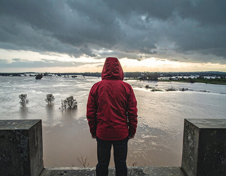 A man in a red jacket stands on a ledge overlooking a flooded area. The sky is cloudy and the water is murky