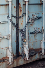 A shipping container stands on a construction site. The container is closed and secured with a chain and padlock. Sunlight shines on the weathered surface.