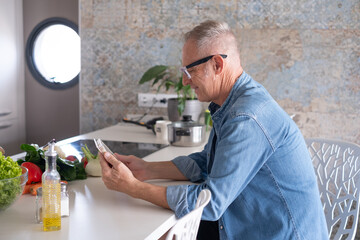 Smiling mature man browsing internet on a digital tablet searching for new recipes before cooking healthy food at home in a modern kitchen, preparing vegetables.
