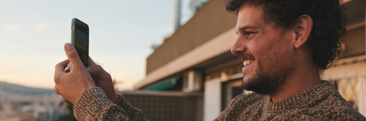 Happy guy stands on balcony at dawn, takes photo, video of beautiful views on smartphone. Panoramic © Andrii Nekrasov