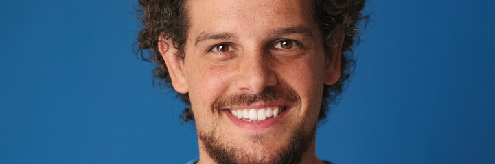 Obraz premium Close-up of guy with curly hair dressed in blue t-shirt looks at the camera with smile on blue background in the studio. Panoramic
