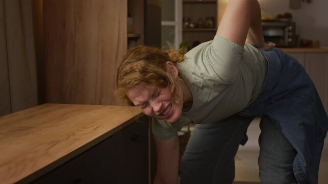 Medium shot of mature Caucasian woman struggling to move heavy drawer chest at home, then spraining lumbar back muscles, grabbing on to waist and crying, unable to get up
