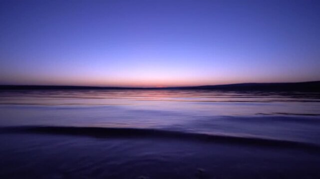 Blurry seascape photograph of a calm ocean at dusk with gentle waves on the shore against a blue and pink gradient sky.