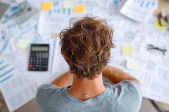 A man with messy hair is sitting at a desk with a calculator, a stack of papers
