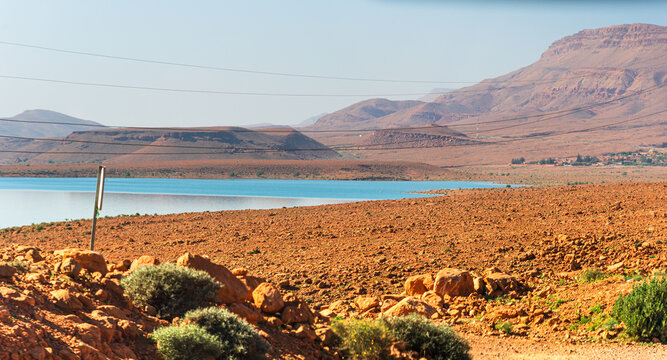 Arid vegetation and sunlit hills meet the blue expanse of Barrage Al-Hassan Addakhil in Parc National Al Hassan Addakhil
