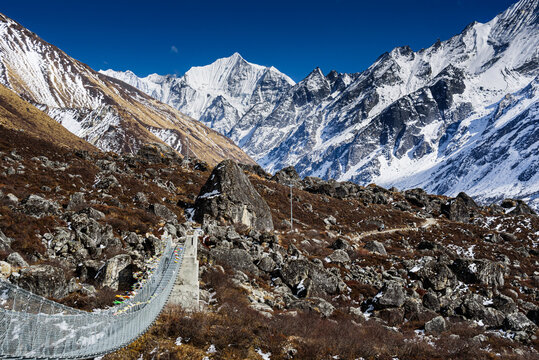 Suspension bridge crossing rocky terrain in Langtang Valley with panoramic views of Gangchempo and surrounding Himalayan peaks under bright blue skies