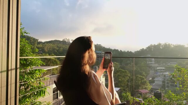 Young woman filming sunrise on smartphone from luxury hotel terrace in Ella, Sri Lanka. Golden morning light and tropical mountain view.