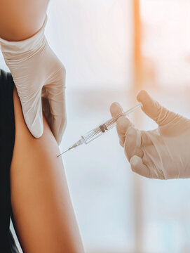 Healthcare worker in gloves gives vaccine injection to patients arm in a clinical environment, with soft-focus background elements visible