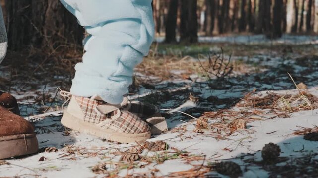 Close-up of toddler legs in blue pants walking on snowy ground with an adult nearby. A concept of first steps, family care, and parental support. Slow motion.