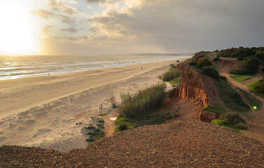 View of empty beach with steep rock cliff, ocean waves, stones, wet golden sand and green vegetation , Portugal.