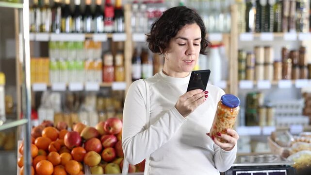 Woman gets acquainted with food scan QR code, take reading canned beans label using phone camera in shop. Client view kidney bean package, inspect manufacturer information using mobile application