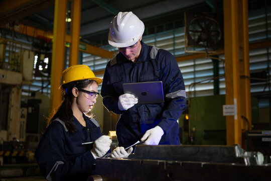 Concentrated male engineer and female technician wearing safety hardhat and glove holding tablet working together inspecting machinery part in metal manufacturing factory