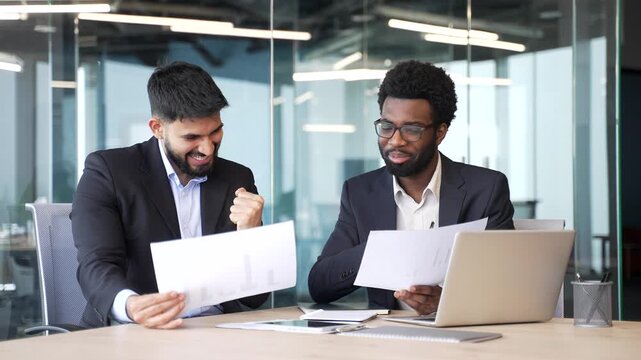 Two businessmen in suits sitting at office desk holding documents and smiling with satisfaction. Colleagues celebrating successful deal, reading positive results and discussing business achievement.