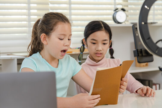 Generation alpha children growing up with education technology, online learning at home, two girls reading book aloud and recording video with smartphone, modern homeschool, digital content creation
