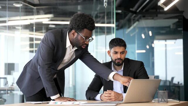 Businessman standing at desk advising and explaining project on laptop computer to seated employee. Male manager mentoring colleague, discussing strategy and guiding work in modern corporate office.