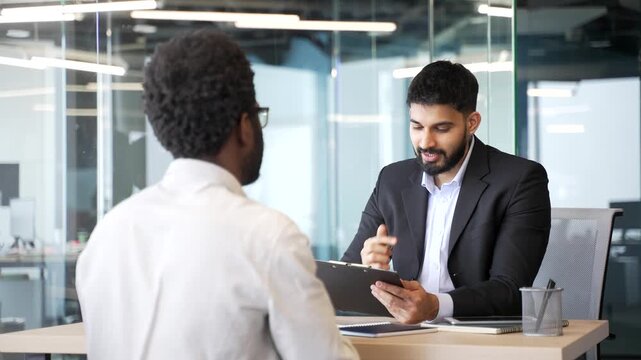 HR manager sitting at office desk talking to candidate during job interview and writing notes on clipboard. Employer evaluating applicant, listening, assessing experience in corporate environment.