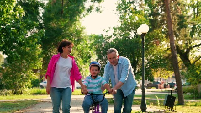Child cyclist with parents in city park. Family day out, father mother teaching son ride bike in park. Happy family, small child in helmet, dad mom, playing on bike outdoors in summer. People in park