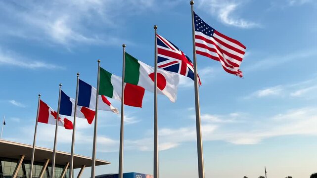 G7 flags waving on poles. A group of G7 national flags wave together on separate flagpoles, aligned in a row and moving in unison under open sky.