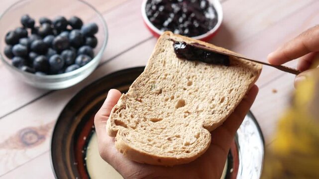 Person spreading organic blueberry jam on slice of whole wheat bread for breakfast