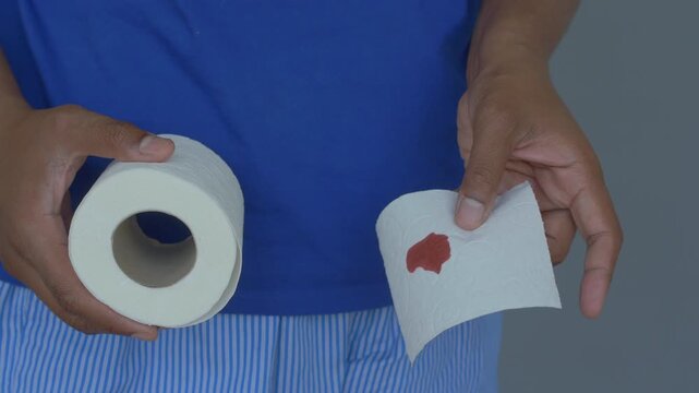A man in blue shirt and striped pajamas holding a toilet paper roll and a piece of toilet paper with red blood stain, indicating a medical concern or health issue